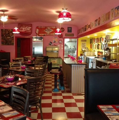 A retro diner with pink walls, checkered floors, and vintage decor, featuring tables and a counter area.