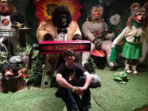 A person sits on the ground in front of colorful animatronic animals playing instruments in a whimsical setting.