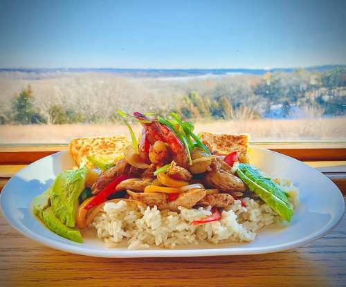 A plate of stir-fried chicken and vegetables on rice, with toast, set against a scenic view of hills and sky.