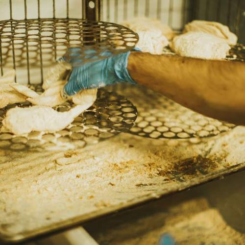 A gloved hand places breaded chicken pieces onto a perforated tray in a kitchen setting.