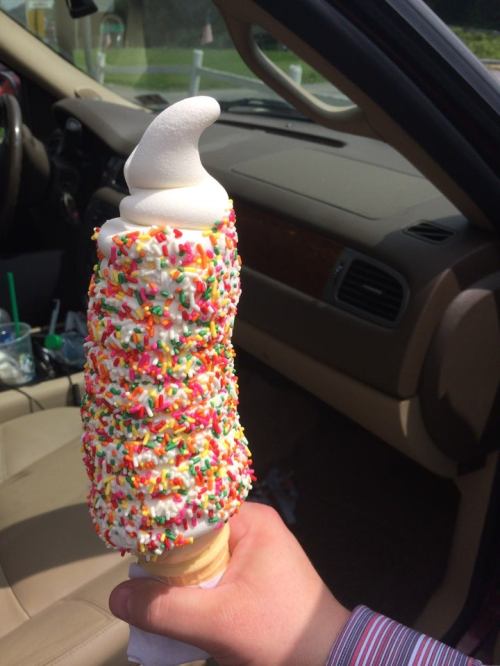 A hand holding a large ice cream cone topped with soft serve and covered in colorful sprinkles, inside a car.