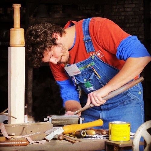 A man in overalls works intently on a wooden project, surrounded by tools in a workshop.
