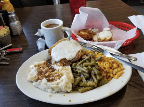A plate of fried chicken with gravy, mashed potatoes, green beans, and corn, alongside coffee and a biscuit.