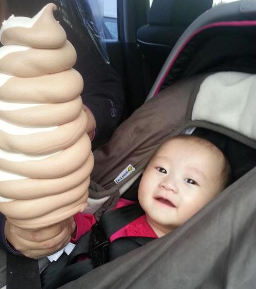 A baby in a car seat smiles at a person holding a large soft-serve ice cream cone.