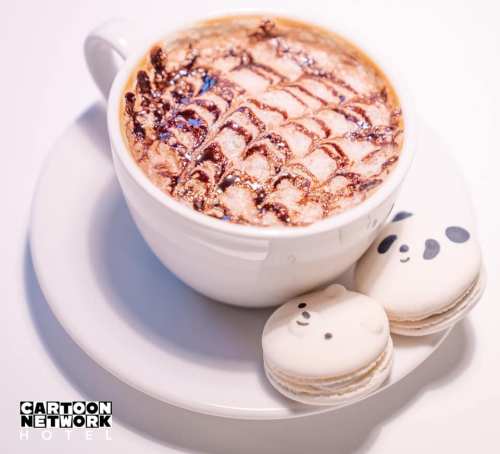 A cup of coffee with intricate chocolate art, accompanied by two cute panda-themed macarons.