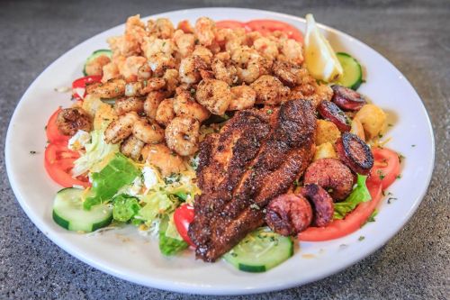 A colorful plate featuring seasoned shrimp, fish, sausage, lettuce, tomatoes, and cucumbers, garnished with lemon.