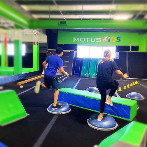 Two kids navigating an indoor obstacle course with balance beams and inflatable platforms in a vibrant gym setting.