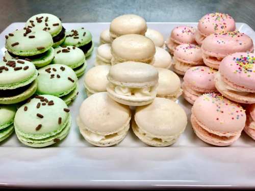 A platter of colorful macarons: green with chocolate sprinkles, white, and pink with sprinkles, arranged neatly.