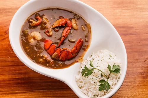 A bowl of seafood gumbo with a large lobster claw, served alongside a portion of white rice and a sprig of parsley.