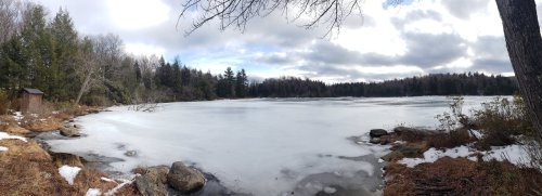 A serene winter landscape featuring a frozen lake surrounded by trees and rocky shores under a cloudy sky.