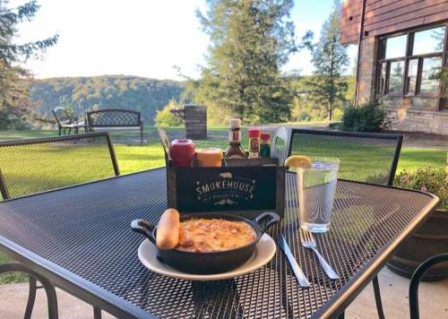 A table set outdoors with a skillet dish, condiments, and a glass of water, surrounded by a scenic landscape.