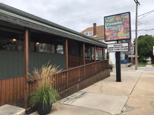 Exterior view of Coombs restaurant featuring a sign with daily specials and a wooden deck with decorative plants.