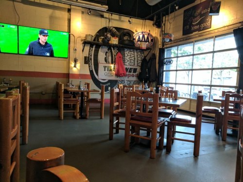 A casual restaurant interior with wooden tables, chairs, and a TV showing a sports game. Large windows let in natural light.