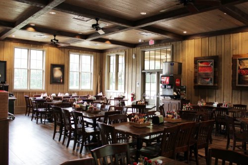Interior of a rustic restaurant with wooden tables, chairs, and large windows, featuring a soda machine and condiments on tables.
