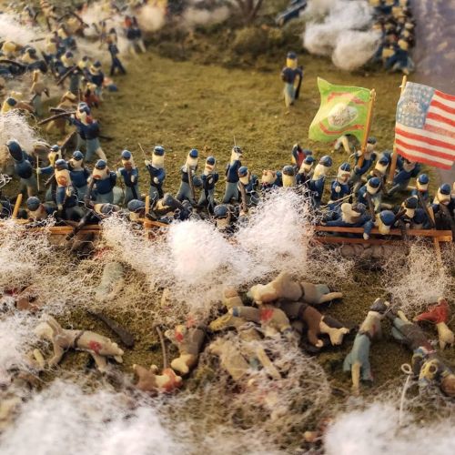 A miniature battlefield scene with toy soldiers, flags, and scattered figures on a grassy terrain.