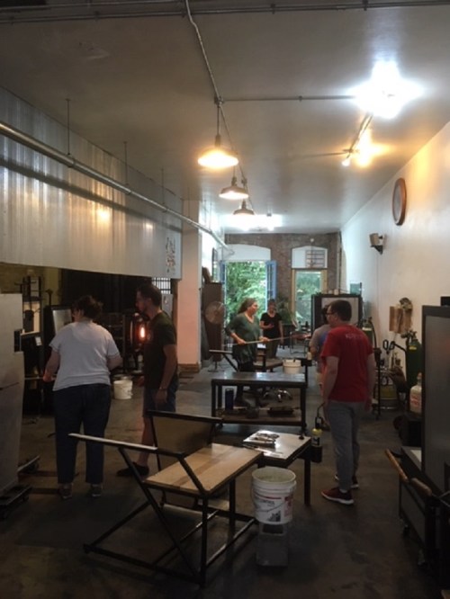 A glassblowing studio with several people working on projects, surrounded by tools and furnaces. Bright and spacious interior.