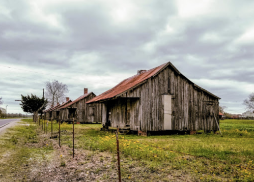 Abandoned wooden houses with rusty roofs beside a road, set against a cloudy sky and grassy field.