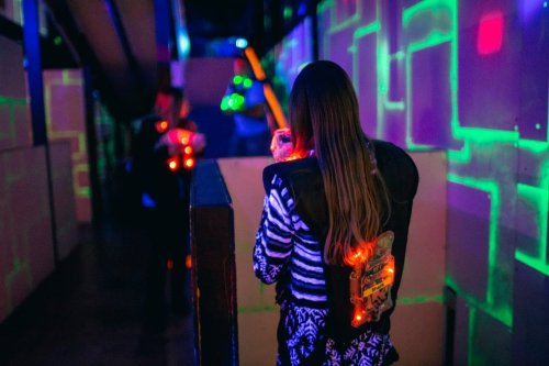 A person in a laser tag arena, wearing a vest with lights, aims at opponents in a colorful, neon-lit environment.