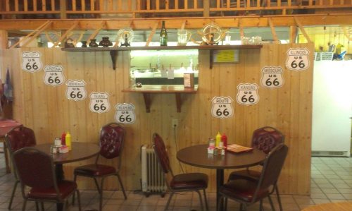 A rustic diner interior with wooden walls, Route 66 signs, and two tables with red chairs and condiments.