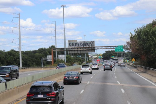 Traffic on a highway with a large overhead sign and trees in the background under a partly cloudy sky.