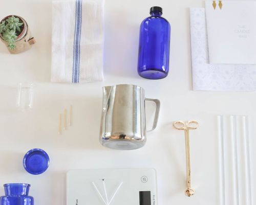 Flat lay of candle-making supplies: blue bottles, a metal pitcher, a scale, wooden sticks, and a towel.