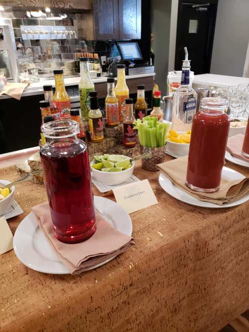 A table with two glasses of cranberry juice, garnished with lime and celery, surrounded by various drink mixers and condiments.