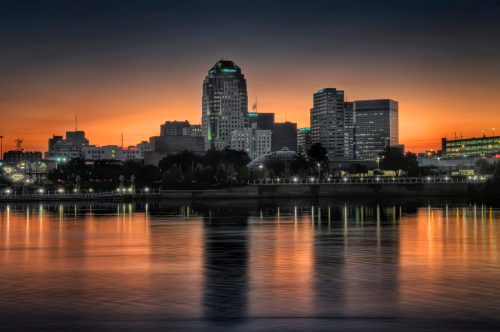 City skyline at dusk, reflecting on water, with buildings illuminated against a vibrant orange and blue sky.