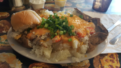 A loaded baked potato topped with cheese, green onions, and seafood, served with a roll on a colorful table.