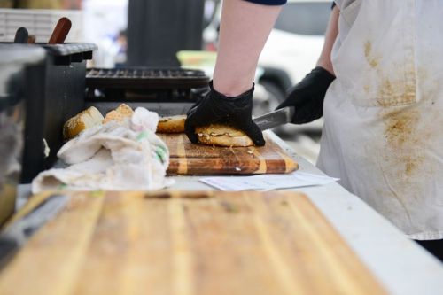 A person in gloves slices bread on a wooden cutting board at a food preparation station.