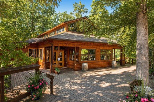 A rustic wooden cabin surrounded by trees, featuring large windows and a welcoming porch with flower planters.