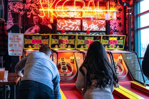 Two people play skee-ball at a vibrant arcade, with neon lights and colorful game machines in the background.