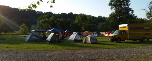 A campsite with several tents, a blue canopy, and a yellow moving truck in a green, wooded area.