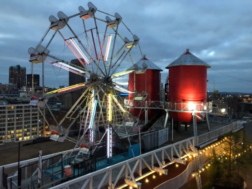 A rooftop scene featuring a colorful Ferris wheel and two red silos against a twilight city skyline.