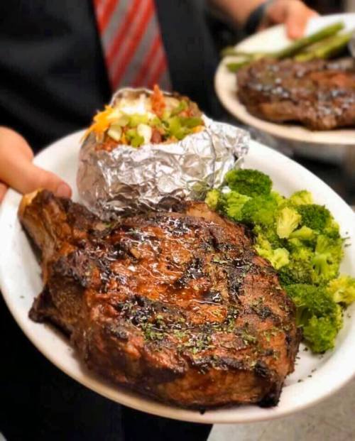 A person holds a plate with a grilled steak, a baked potato topped with vegetables, and a side of broccoli.