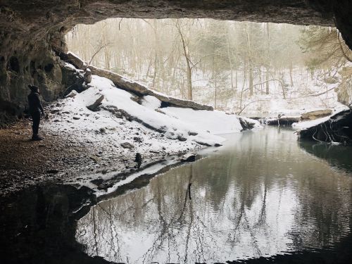 A person stands inside a cave, looking out at a snowy landscape and a calm, reflective body of water.