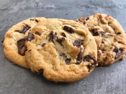 Three freshly baked chocolate chip cookies on a dark stone surface.