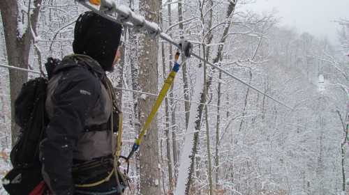 A person in winter gear stands on a snowy zip line platform, surrounded by snow-covered trees.