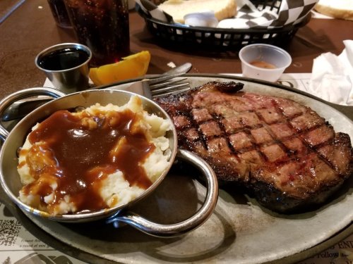 A grilled steak with grill marks, served with mashed potatoes and gravy, alongside a drink and a slice of orange.