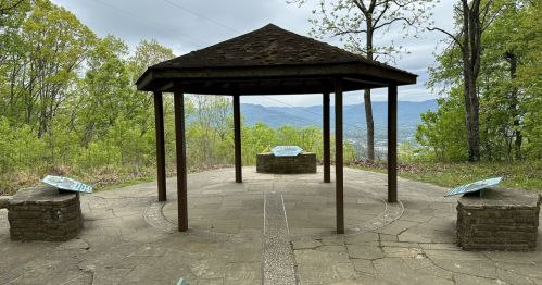 A gazebo with stone benches overlooks a scenic view of mountains and greenery under a cloudy sky.