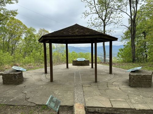 A gazebo on a hillside surrounded by trees, with stone tables and scenic mountain views in the background.