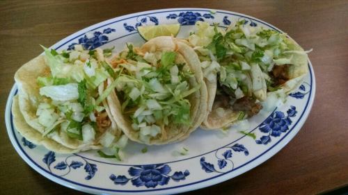Three tacos on a decorative plate, topped with chopped onions and cilantro, with a lime wedge on the side.