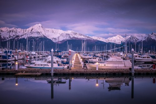 A serene marina at dusk, with boats docked and snow-capped mountains reflecting in calm waters.