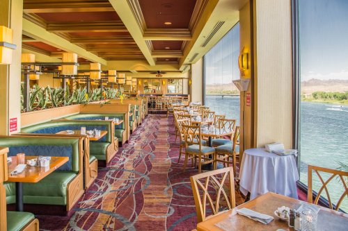 Brightly lit restaurant interior with green booths and tables, overlooking a scenic view of water and greenery.