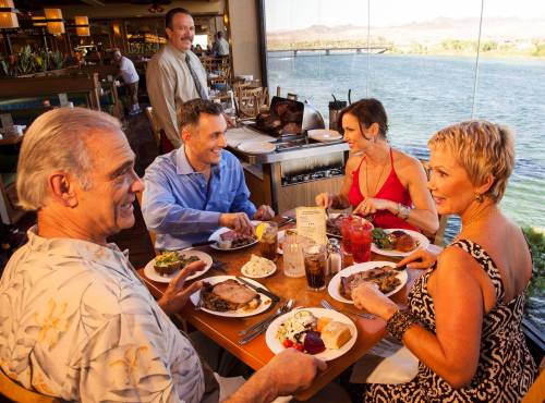 A group of five people enjoying a meal together at a restaurant with a scenic view of water and mountains.