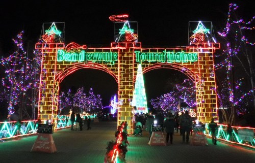 Colorful holiday lights illuminate an archway reading "Bentonville Tour of Lights," with festive decorations and trees.