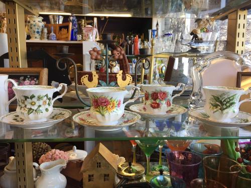 Three decorative teacups with floral designs displayed on a glass shelf, surrounded by various collectibles.