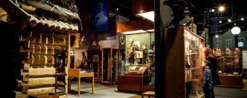 A museum interior featuring a log cabin, display cases with artifacts, and visitors exploring the exhibits.