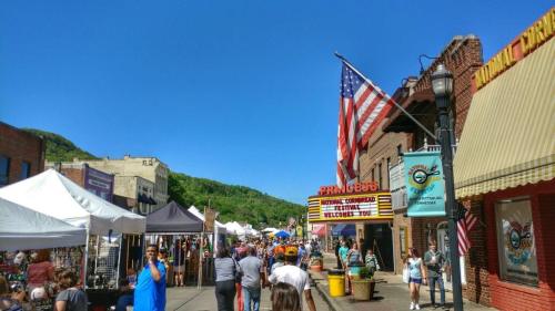A bustling street fair with tents, people, and an American flag against a clear blue sky.