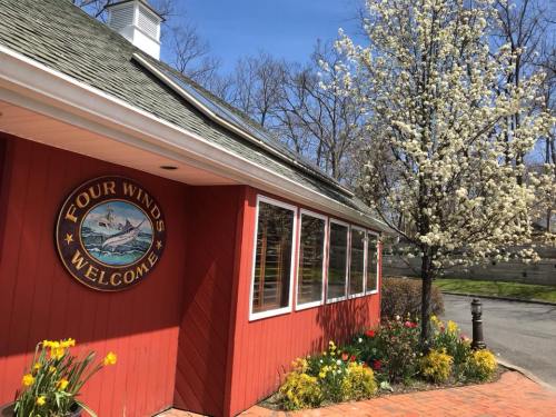A red building with a "Four Winds Welcome" sign, surrounded by blooming flowers and a tree on a sunny day.