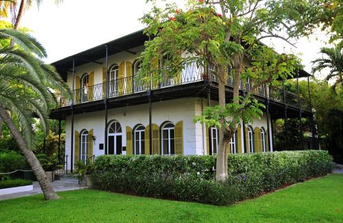 A two-story white house with yellow shutters, surrounded by lush greenery and palm trees.